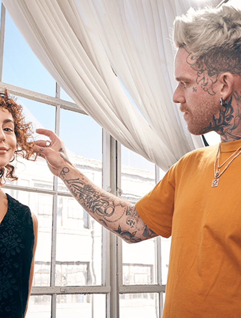Male barber with short blonde hair and tattoos dressed in an orange shirt styling model's hair